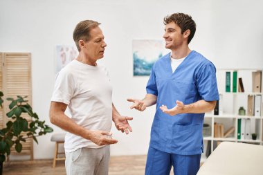 joyous handsome doctor in blue robe talking to his mature patient during appointment in hospital clipart