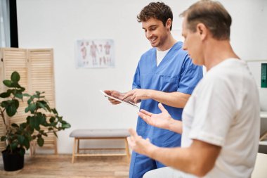 Mavi bornozlu yakışıklı doktor elinde tabletle hastanedeki olgun hastayı dinliyor.