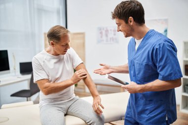 Mavi bornozlu neşeli çekici doktor elinde tabletle hastanedeki olgun hastasını dinliyor.