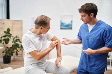 İyi görünümlü, çalışkan bir doktor elinde tabletle yetişkin hastasının rehabilitasyonunu dinliyor.