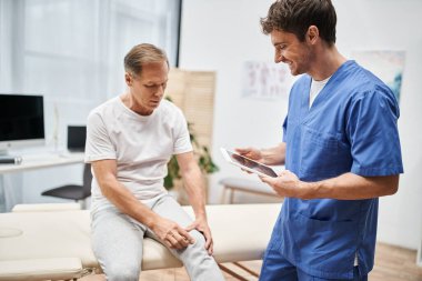 Mavi bornozlu neşeli doktor elinde tabletle hastanedeki olgun hastasını dinliyor.