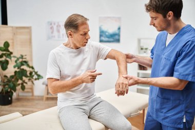 Hastanedeki rehabilitasyon sırasında yetişkin hastasıyla çalışan mavi cüppeli yakışıklı bir doktor.