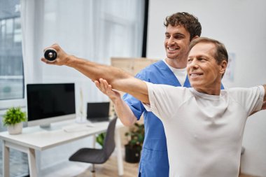 Mavi cüppeli neşeli doktor rehabilitasyon sırasında halterli hastasına yardım ediyor.