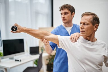 Mavi bornozlu çalışkan doktor rehabilitasyon sırasında halterli olgun hastasına yardım ediyor.