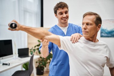 Mavi bornozlu neşeli doktor rehabilitasyon sırasında halterini kullanan yakışıklı hastasına yardım ediyor.