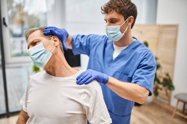 Maskeli ve eldivenli yakışıklı doktor rehabilitasyon sırasında olgun hastasını geriyor.