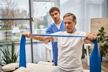 hardworking doctor in blue uniform helping his mature patient with resistance band on rehabilitation clipart