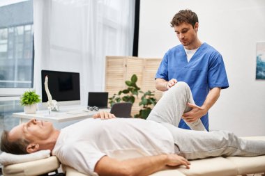 Mavi cüppeli yakışıklı doktor, yetişkin hastasının rehabilitasyon sırasında esnemesine yardım ediyor.