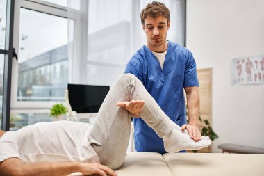 handsome hardworking doctor in blue robe helping his mature patient to rehabilitate in hospital clipart