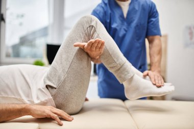 cropped view of hardworking doctor in blue robe stretching his mature patient in hospital ward clipart