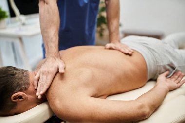 cropped view of hardworking doctor in blue robe massaging his mature patient during rehabilitation clipart