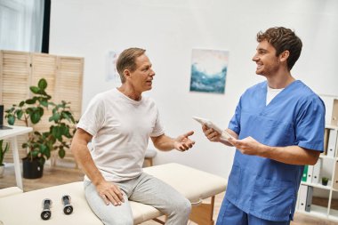 Rehabilitasyon sırasında olgun, neşeli hastasıyla konuşan tabletli mutlu çalışkan bir doktor.