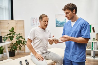 Mavi cüppeli yakışıklı doktor elinde tabletle randevusundaki olgun hastasını dinliyor.