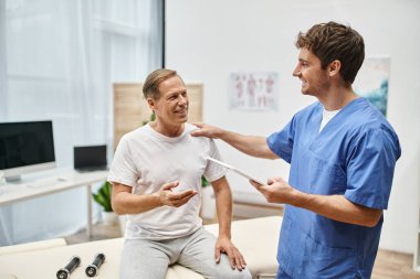 jolly dedicated doctor holding tablet and talking to his mature merry patient during rehabilitation clipart