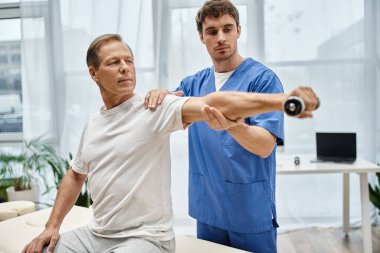 Yakışıklı doktor, yetişkin hastasına günlük giysiler içinde rehabilitasyon için dambıl kullanmasında yardım ediyor.