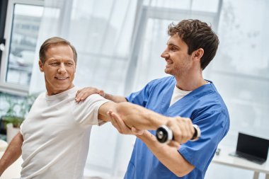 Yakışıklı, neşeli, neşeli, olgun hastasına rehabilitasyon sırasında halter kullanması için yardım eden doktor.
