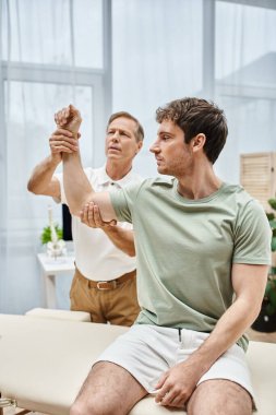 Üniformalı iyi görünümlü bir rehabilitasyon uzmanı randevusu sırasında hastanın esnemesine yardım ediyor.