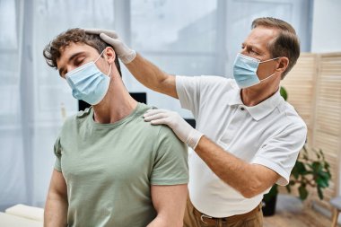 Maskeli ve eldivenli iyi görünümlü olgun bir doktor hastasının hastanede iyileşmesine yardım ediyor.