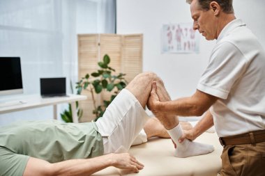 good looking mature doctor checking up knees of his patient in hospital, rehabilitation concept clipart