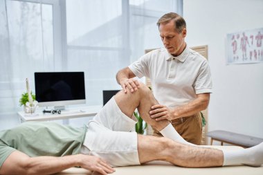 İyi görünümlü olgun bir doktor hastasının dizlerini kontrol ediyor, rehabilitasyon konsepti.
