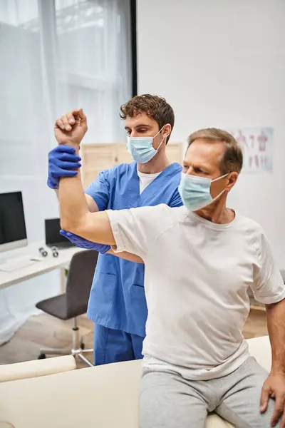 stock image hardworking doctor with mask and gloves helping mature patient to rehabilitate his muscles in ward