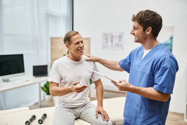 Stock image jolly dedicated doctor holding tablet and talking to his mature merry patient during rehabilitation