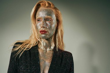 Parlak elbiseli, parlak sarışın kadın vücudunun her yerinde parıltı ve yüzü gri arkaplanda poz veriyor.
