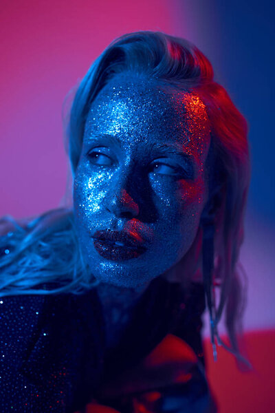 portrait of pretty blonde woman with glitter on face and shiny outfit posing on neon background