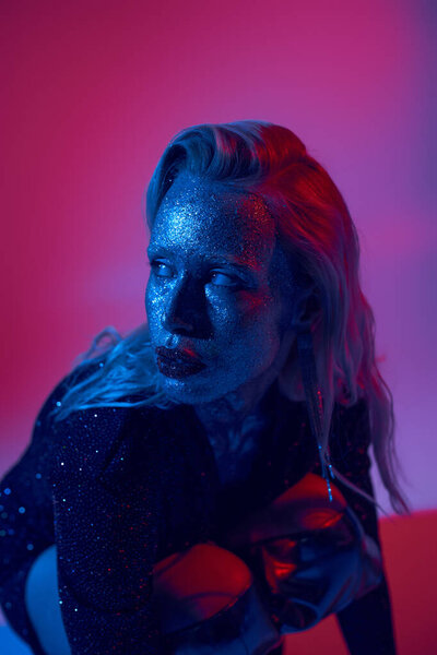 portrait of attractive blonde woman with glitter on face and shiny outfit posing on neon background