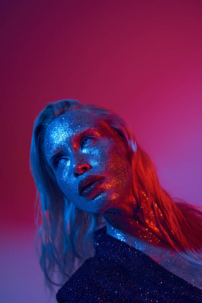 blonde and shiny woman with glitter on face and sparkling outfit posing on vibrant neon background