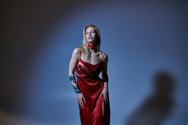 alluring fashionable woman in red chic dress with tattoo posing and looking away on dark backdrop