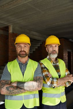 two good looking bearded men in safety vests and helmets looking at camera, cottage builders clipart