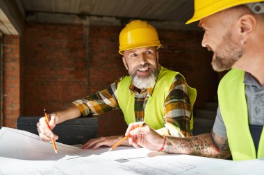cheerful good looking builders in safety vests and helmets looking at blueprint of building clipart
