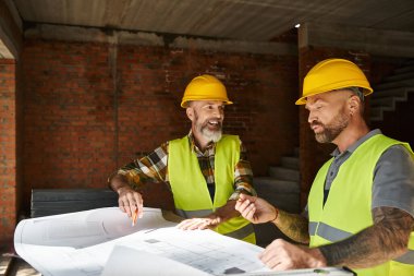 joyous handsome builders in safety vests and helmets looking at blueprint before construction clipart