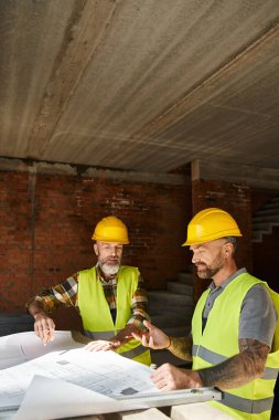 two handsome builders in safety helmets and vests working with blueprint before construction clipart