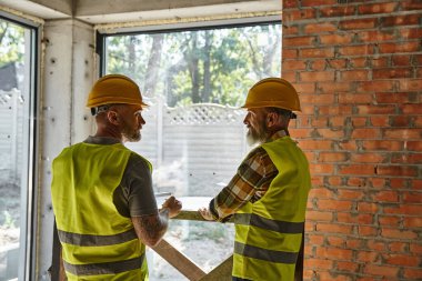 two hardworking bearded men in safety vests and helmets on construction site, cottage builders clipart
