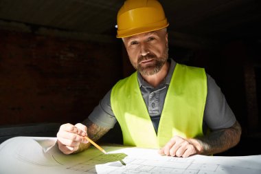 handsome builder in safety vest working on his blueprint on construction site and looking at camera clipart