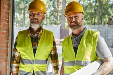 good looking dedicated builders in safety vests and helmets looking at camera on construction site clipart