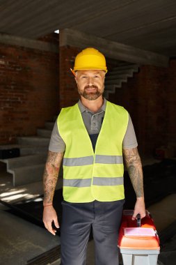 cheerful good looking builder in safety vest and helmet holding toolbox and looking at camera clipart