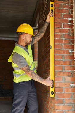 Güvenlik yeleği giymiş, sakallı yakışıklı inşaat işçisi tuğla duvarı seviyesiyle ölçüyor, inşaatçı