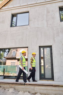 Güvenlik kaskı takmış yakışıklı kır evi inşaatçıları. Alet kutusu seviyesinde ve bina planıyla dışarıda çalışıyorlar.