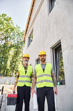 good looking builders with toolbox level and blueprint looking at camera while working outside clipart