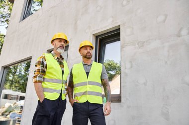 appealing devoted builders in safety helmets and vests posing outside on construction site clipart