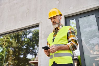 Çelik yelek giyen düşünceli yakışıklı adam inşaat sahasında, yazlık inşaatçıyken telefonuna bakıyor.