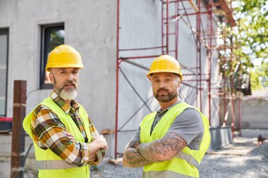 good looking cottage builders in safety helmets posing next to scaffolding and looking at camera clipart