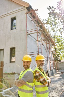 Güvenlik yelekli neşeli yazlık inşaatçılar iskelenin yanında sırt sırta poz verip kameraya bakıyorlar.