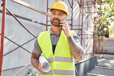 Güvenlik yeleğinde dövmeleri olan yakışıklı sakallı bir yazlık inşaatçı elinde planlar ve telefonla konuşuyor.