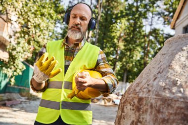 Güvenlik eldiveni ve yelek giyen, kulaklık ve kaskla poz veren çalışkan bir yazlık inşaatçısı.