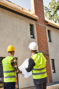 Kır evi inşaatçısı ve mimarı ellerinde planlar ve inşaat alanlarını tartışıyorlar.