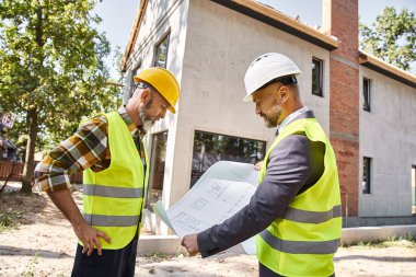 Kır evi inşaatçısı ve mimarı ellerinde planlar ve inşaat alanlarını tartışıyorlar.
